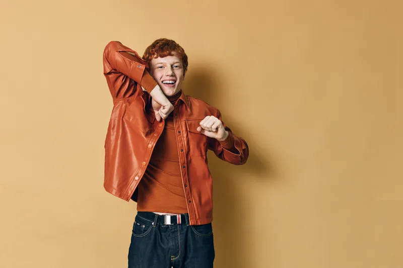 Happy young man with red hair wearing orange jacket and sweater dancing and smiling against beige background in casual style with jeans and belt.