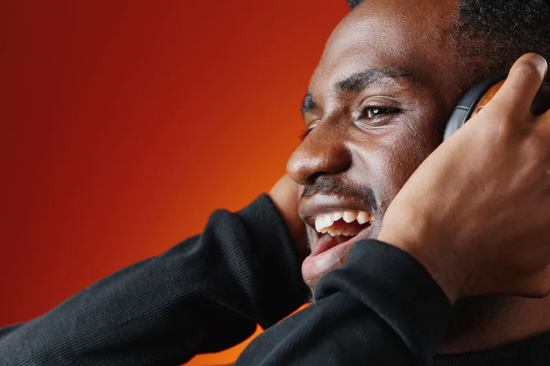 Happy African man enjoying music with headphones, against a vibrant orange background, expressing joy and excitement in a stylish black outfit