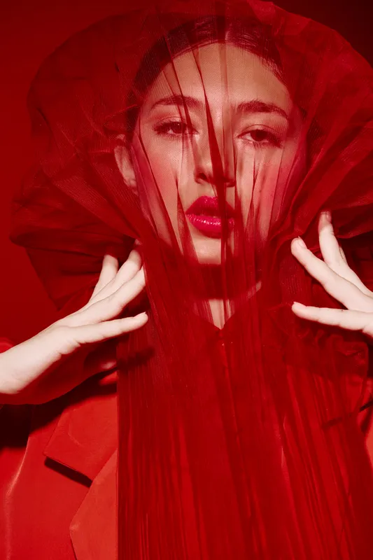 Portrait of woman with red tulle fabric covering her face, wearing red lipstick and coat. Dramatic fashion and beauty concept with creative styling in a studio setting.