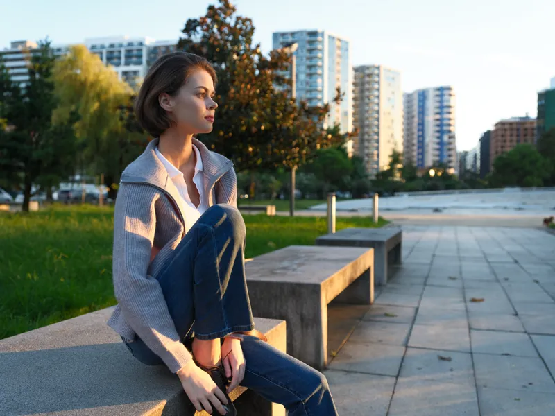 A thoughtful woman sits on a concrete bench in an urban park during golden hour, reflecting serenity and focus amid a modern cityscape and green surroundings.