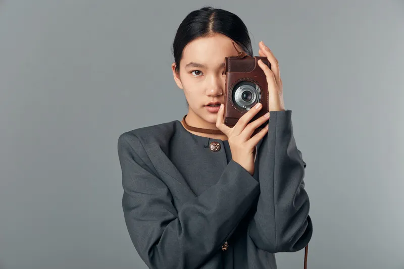 Young woman in gray suit holding vintage camera close to face against neutral background. Portrait with calm expression and photography equipment for creative concept and hobby.
