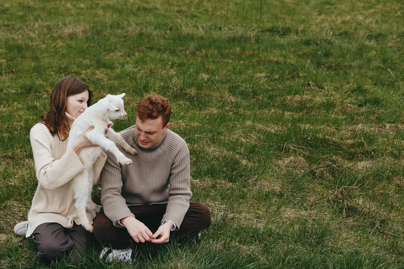 A couple sits on grass outdoors, holding a small white goat kid between them, smiling and relaxed. A lifestyle moment of gentle bonding, nature, and shared care in a casual, sunny setting.