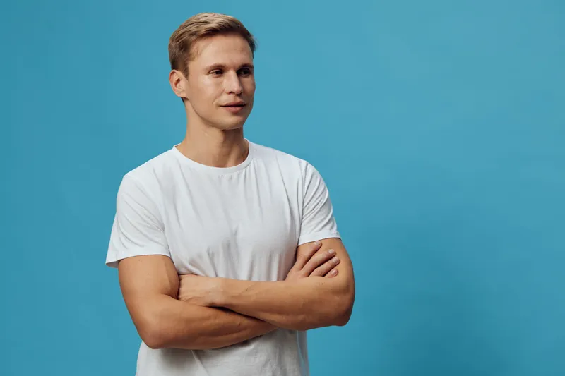 Full body portrait of a young man with light brown hair wearing a plain white t-shirt, standing with arms crossed against a solid blue background. Casual fashion concept, youthful style, relaxed mood,