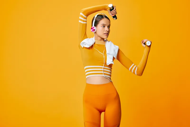 Fitness scene with a athletic woman in bright orange sportswear performing a dynamic workout, lifting dumbbells, headphones on, towel over shoulder, vibrant studio backdrop.