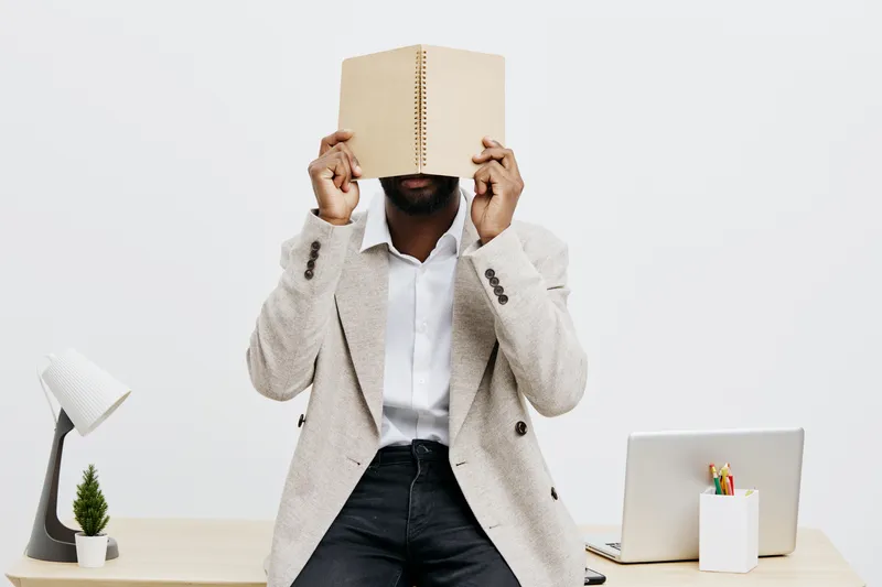 Professional male in casual blazer holding closed book in front of face, sitting at tidy modern office desk with laptop, lamp, plant and stationery, minimal background. Concept: education, reading, wo