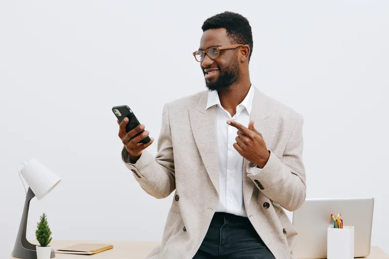 Confident young man with beard and glasses in light grey blazer and white shirt using smartphone, smiling happily in modern office environment with minimal decor and white background, promoting techno