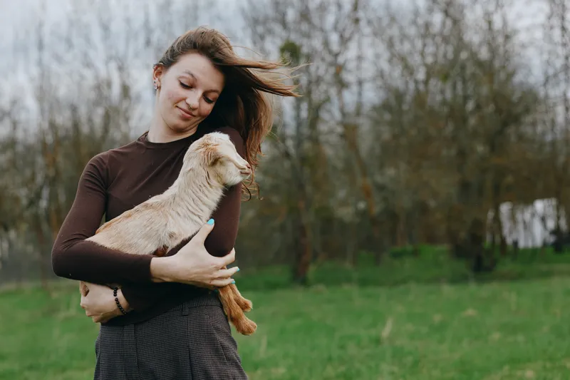 A woman in a brown long sleeve top gently hugs a goat kid in an open field, wind in her hair, smiling softly. A warm, candid lifestyle moment of bonding, care, and rural nature.