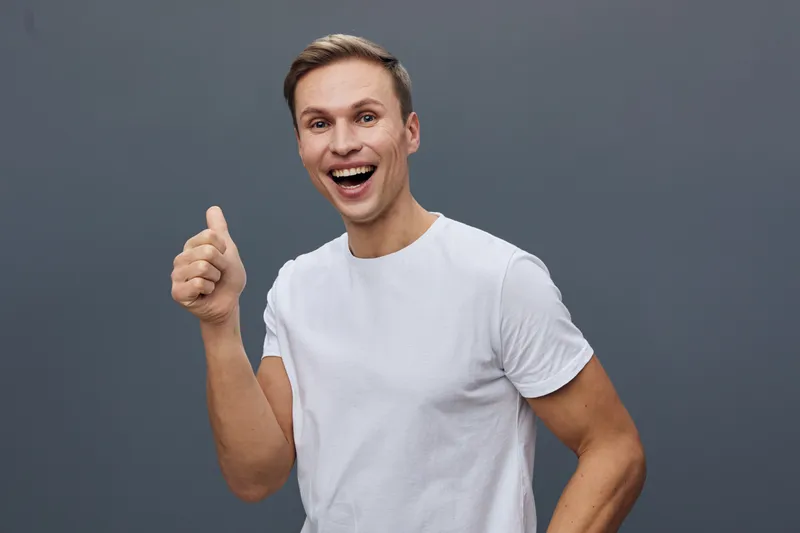 Smiling happy young Caucasian man in casual white t-shirt giving thumbs up gesture isolated on plain gray background studio portrait. People lifestyle concept, positive emotion, cheerful mood, friendl