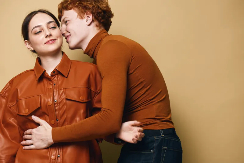 Couple embracing indoors with man kissing woman on cheek, wearing casual autumn clothes in warm tones against beige background for love and affection theme.