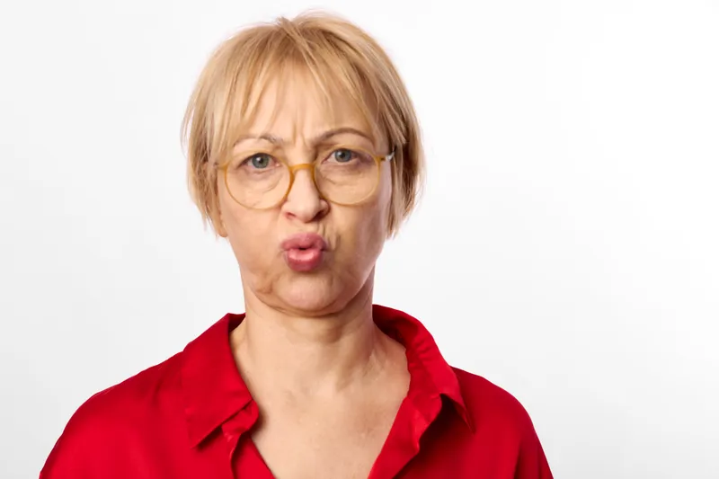 Senior woman with short blond hair and glasses, wearing red polo, surprised expression, tense face, close portrait on white background, emotion of shock and worry