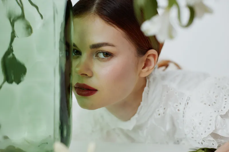 A serene model with pale skin peering from behind a green vase, her expression contemplative and soft, in a minimalistic floral setting