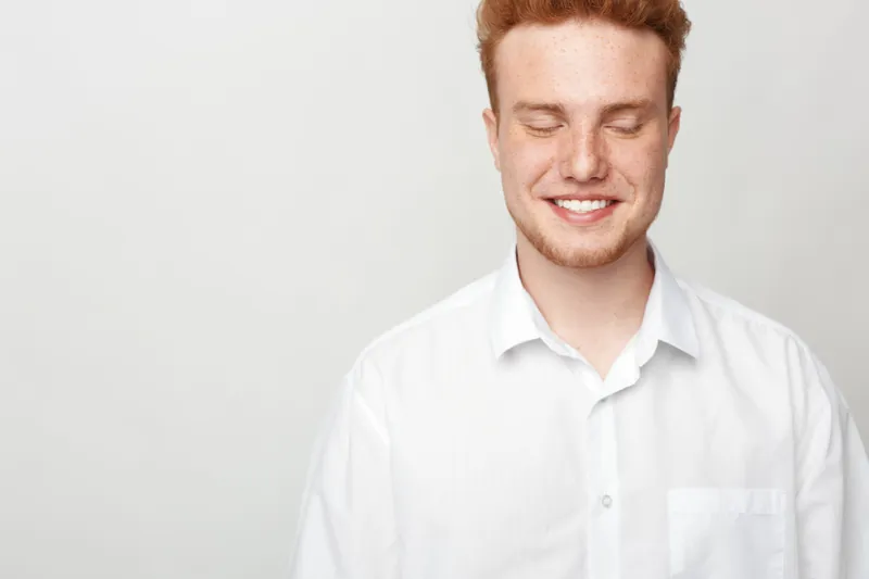 Smiling young man with red hair and closed eyes wears white shirt against plain background. Concept of happiness, confidence, and natural positive emotions in casual portrait.