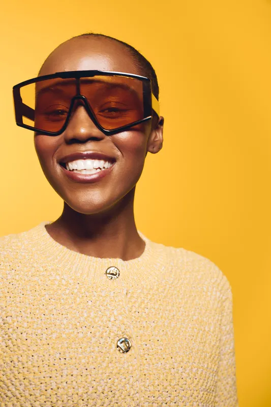 Smiling black African woman with dark skin wearing oversized brown sunglasses and yellow knitted sweater poses against bright yellow background. Concept of fashion, style, and beauty.