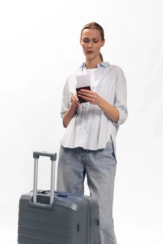 Young woman stands with a suitcase holding a passport and phone, preparing for travel or waiting for a flight. Minimalist white background highlights her focused expression.