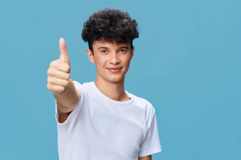 young man with curly hair in white t-shirt showing thumbs up gesture against blue background, positive expression, casual style, studio portrait
