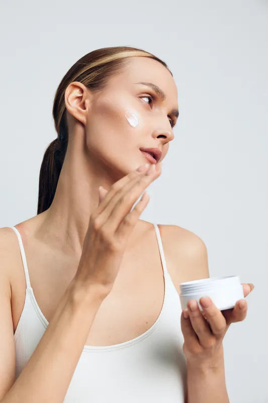 Young woman with brunette hair applies cream on her cheek in a bright studio, capturing a calm skincare moment with a focused expression, soft glow on skin, minimalist background.