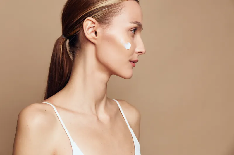 Young woman with clear skin applying facial cream on cheek, side profile portrait, natural light, neutral background, skincare routine, minimal makeup, healthy beauty