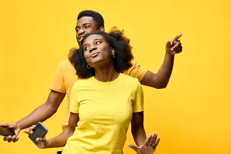 Happy young couple enjoying music together in vibrant yellow outfits, dancing with smartphones against a bright yellow background
