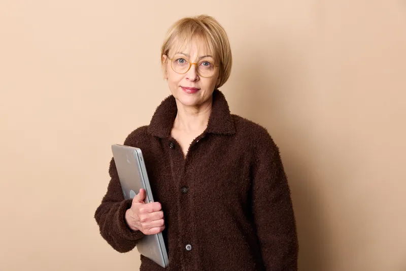 Senior woman with glasses holding a notebook, calm focused expression, wearing a dark coat, studio portrait indoors on beige background, education and learning concept quietly