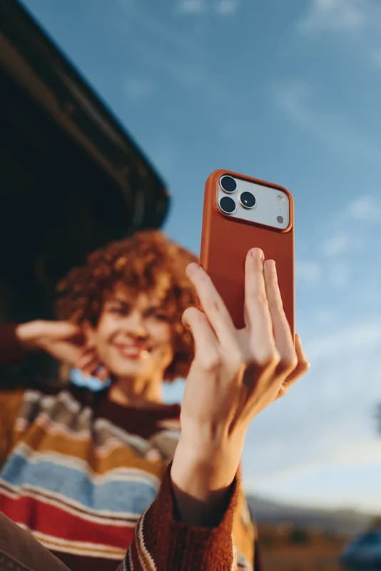 Woman lifestyle selfie smartphone outdoors, casual sweater and curly hair, smiling in warm daylight under blue sky, parked vehicle blurred behind, lifestyle moment