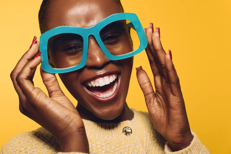 Happy African American woman with dark skin wearing oversized blue glasses and a yellow sweater smiles joyfully against a yellow background. Concept of fashion, happiness, and style.