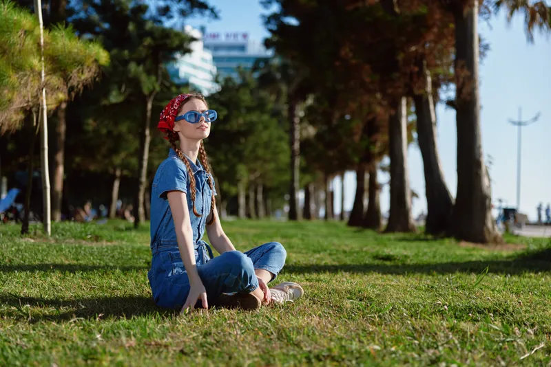 lifestyle woman in modern boho-western denim street style sitting on grass outdoors, film color effect, casual fashion with headband and sunglasses, sunny day