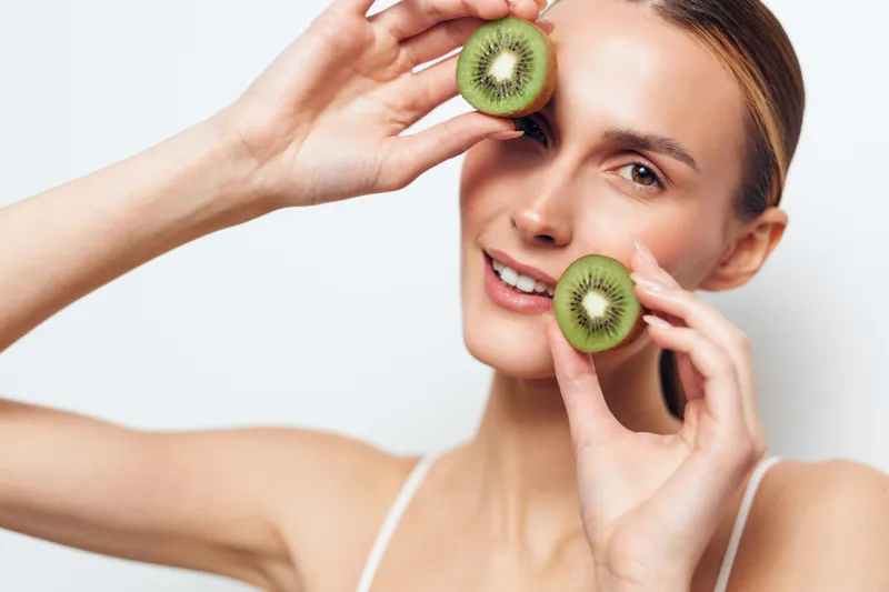 Young woman holds two halves of fresh kiwi fruit near her face smiles naturally isolated on white background healthy lifestyle concept closeup portrait.