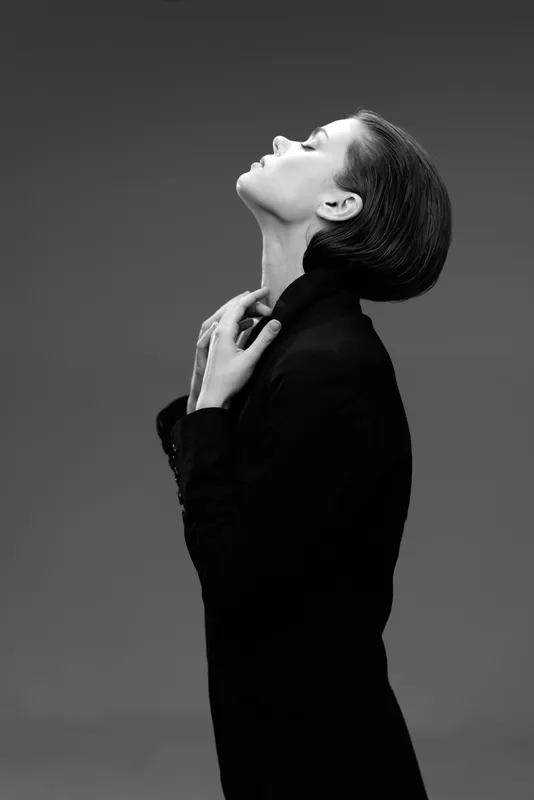 Black and white portrait of a woman in a dark outfit posing with closed eyes and serene expression against grey background.
