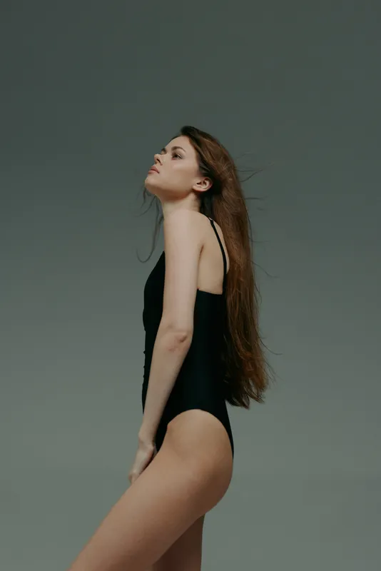 Young woman posing in black swimsuit with long hair against gray background, minimalistic and elegant style.