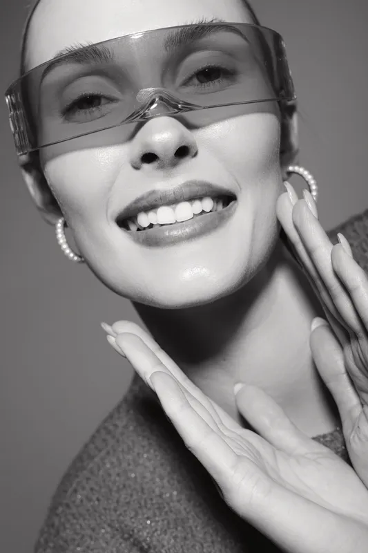 Smiling caucasian woman with fair skin wearing translucent glasses and pearl earrings touches face with hands. Concept of beauty, fashion, and joyful mood.