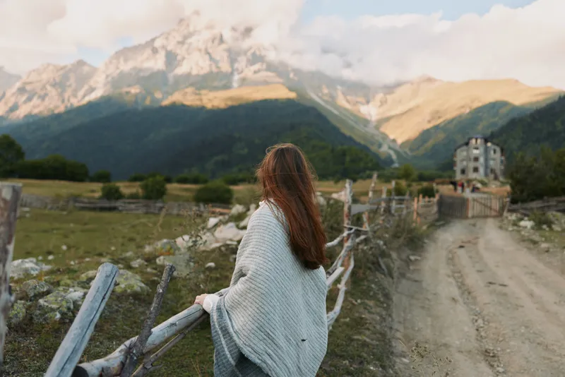 A young woman in a light shawl stands by a wooden fence, gazing toward distant mountains in a quiet rural landscape.
