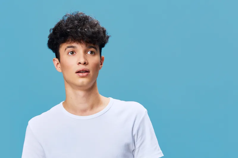 Young man with curly hair wearing a white t-shirt looking surprised against a blue background in a casual contemporary style.