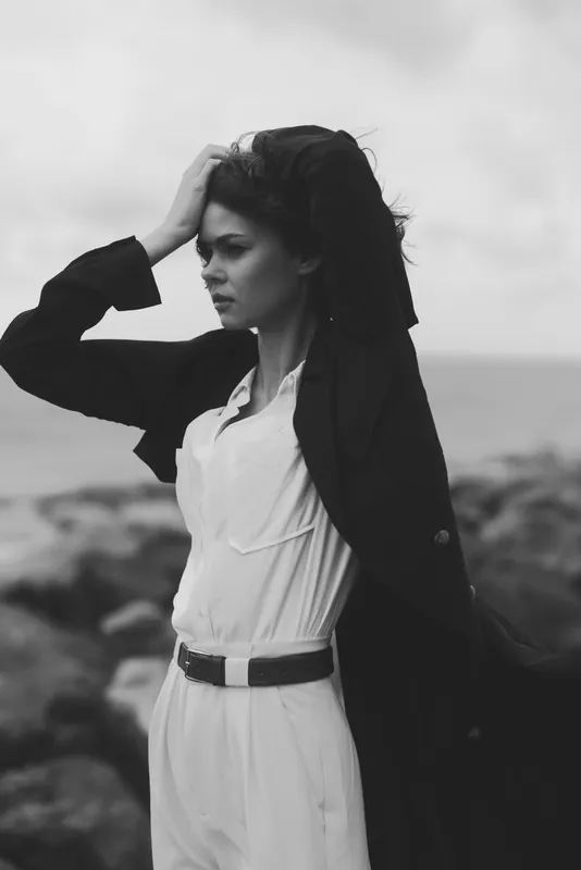Moody outdoor fashion scene with a windblown coat on a woman along the coast, a serene seaside mood under gray skies, captured in monochrome for dramatic contrast and timeless elegance