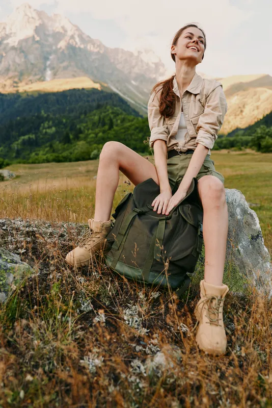 A woman sits on a rock in a sunlit meadow, backpack beside her, smiling with relaxed confidence. Outdoor portrait capturing casual hiking vibe and nature connection.