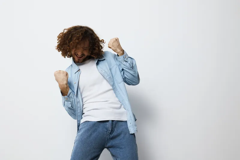 Happy young man celebrating success with joyful gestures, wearing casual denim outfit and showing excitement against a clean white background