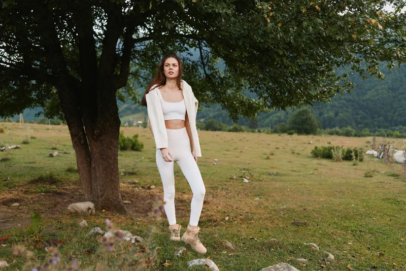Young woman in white outfit stands near tree in green meadow with mountains in background, relaxed lifestyle concept outdoors in natural environment.