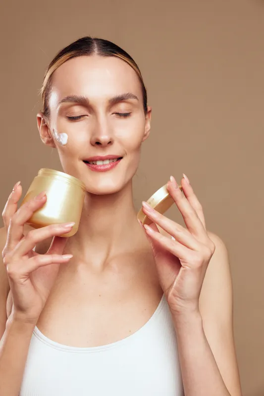 Woman applying skincare cream to her face, self care wellness concept, holding a cosmetic jar and container, beauty closeup portrait, smooth skin, soft studio light, beige background