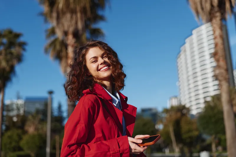 Young woman with curly hair smiles happily while holding a smartphone outdoors on a sunny day with palm trees and modern buildings in the background.