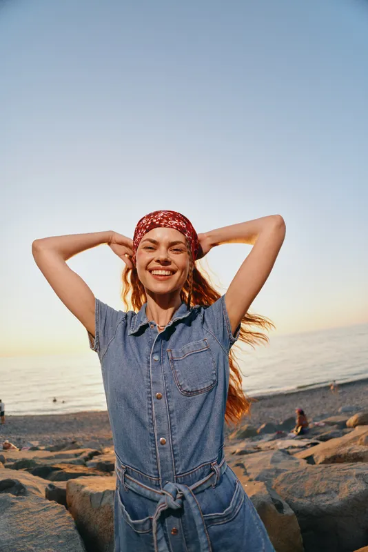 Lifestyle woman in modern boho-western denim street style smiling joyfully at sunset beach with film color tones, casual look, relaxed mood.