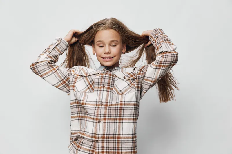 Girl with long hair wearing checkered shirt standing with eyes closed and hands holding hair on light background. Child, casual outfit, youth and playful mood portrait.