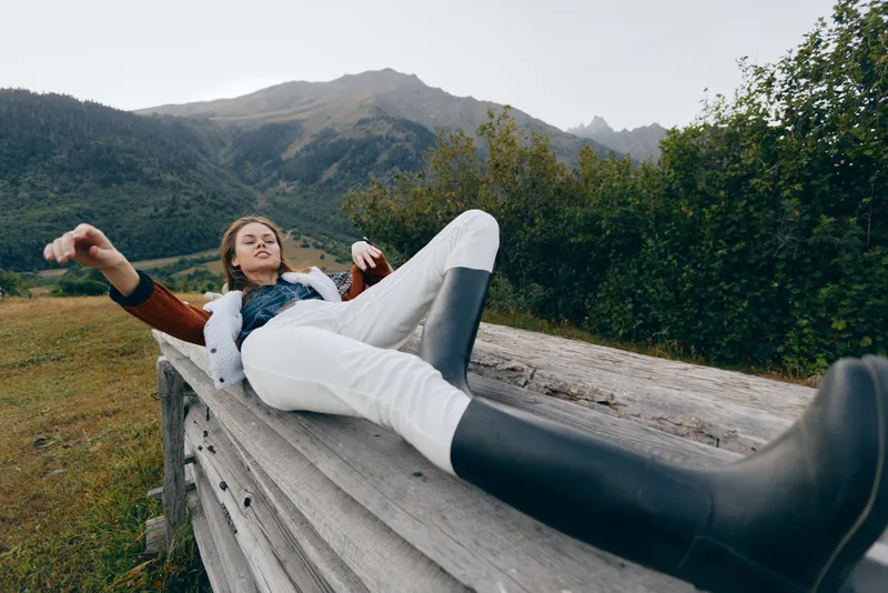 Woman in rubber boots relaxing on a wooden bench in a meadow with mountain backdrop, casual outfit and peaceful nature scene for outdoor leisure, hiking rest and countryside escape