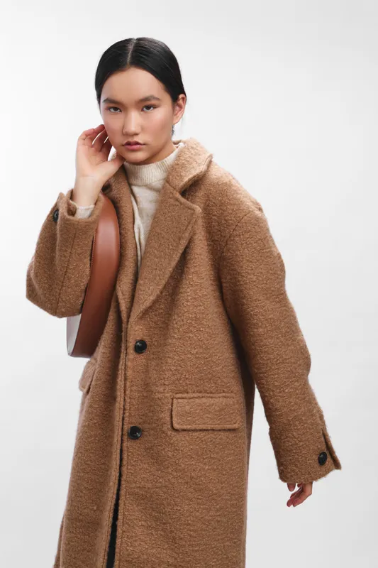 Asian woman wearing a warm beige overcoat stands in a neutral studio, exuding calm confidence and modern chic, a stylish fashion scene with soft lighting.