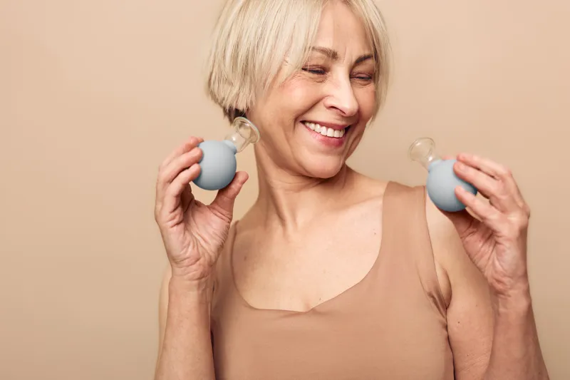 Smiling mature woman with short blonde hair holding silicone suction cups near shoulders in beige top. Concept for alternative therapy, wellness, and self-care for healthy lifestyle.