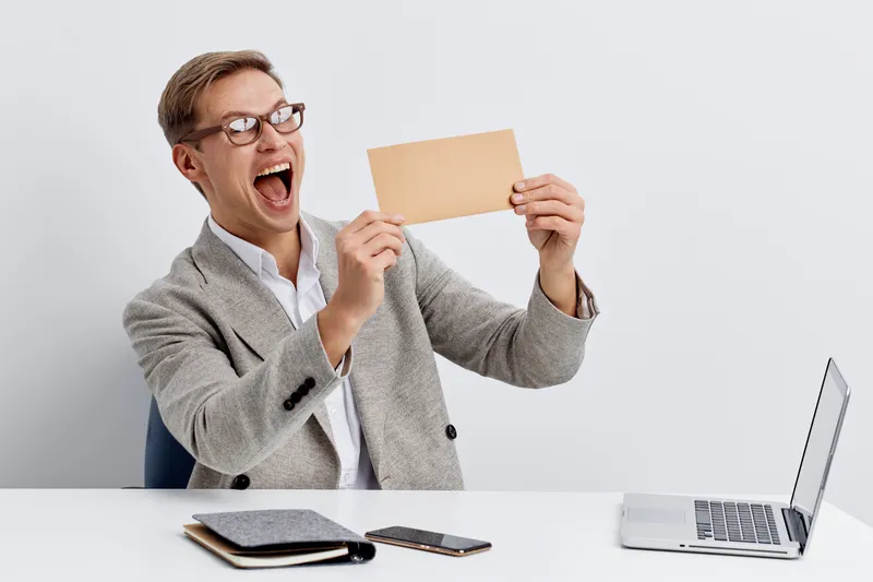 Happy young man with glasses displaying a blank card, sitting at a desk with a laptop, notebook, and calculator on a plain white background. Business, office, excitement, celebration, success, technol
