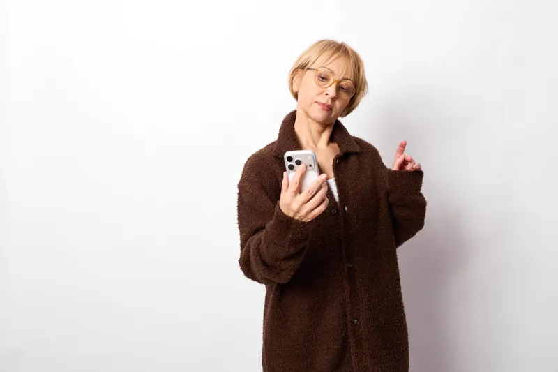 elderly woman smartphone holding portrait glasses coat gesture in white studio, looking at screen with thoughtful expression, warm brown winter outfit, soft even light