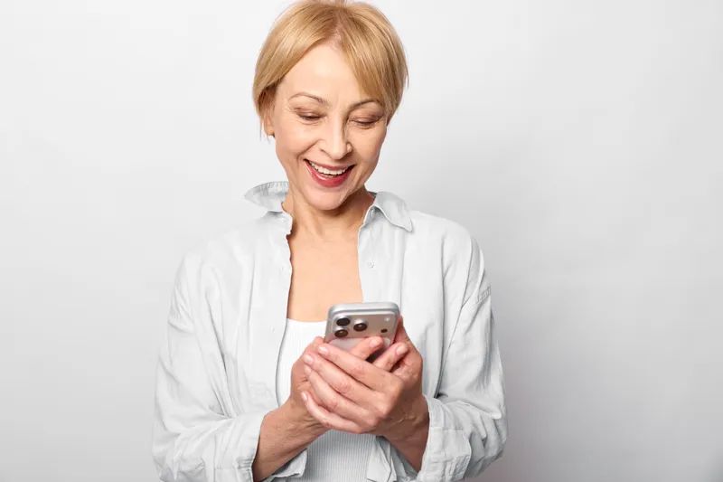 Woman elderly smiling, holding smartphone in hands, using mobile technology for communication, studio portrait on white background, soft light, relaxed pose