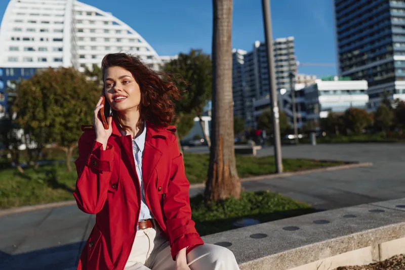 Young woman in red coat talks on smartphone outdoors in city park on sunny day, smiling and enjoying fresh air with modern buildings in background.