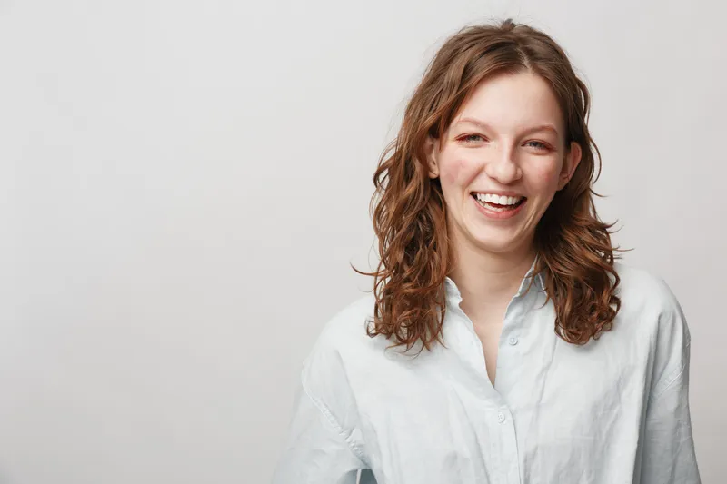 Smiling young woman with curly hair wearing white shirt looks happy and confident. Studio portrait on light background. Concept of positive emotions, natural beauty, and casual style.