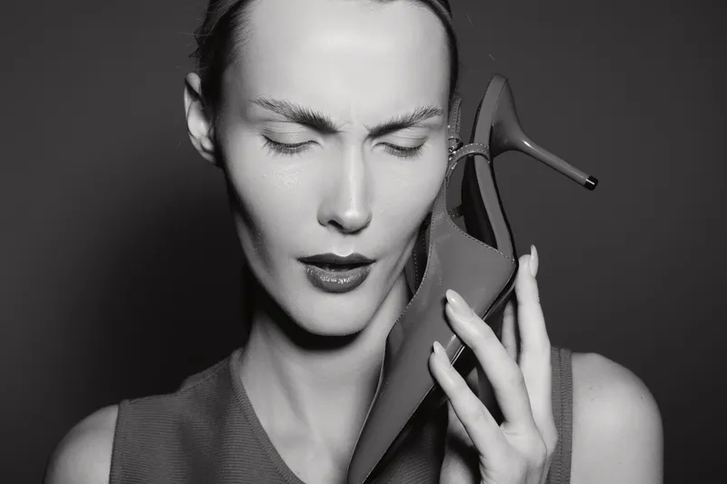 Closeup of a stressed white woman with pale skin holding a high heel shoe to her ear, eyes closed, expressing frustration in black and white studio lighting.