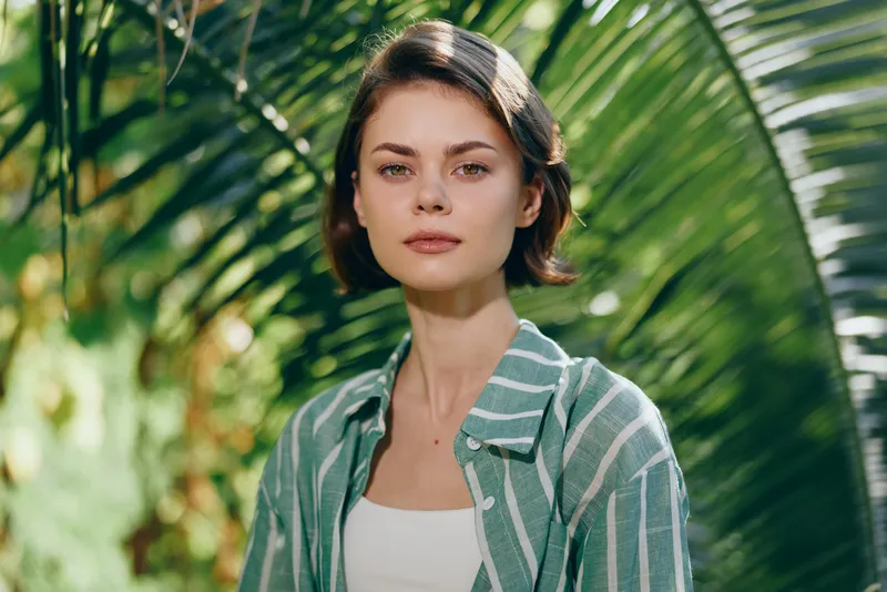 Portrait of a young woman outdoors under tropical light, wearing a green striped shirt over a white top, with a calm, focused expression.